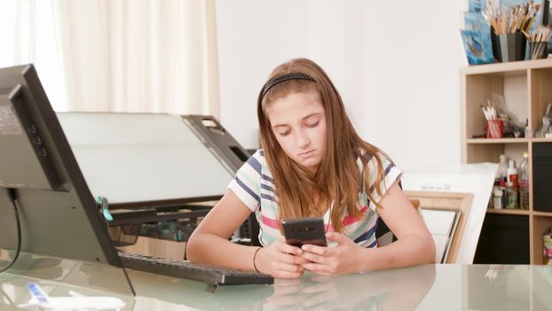 Bored teenage girl sits at a desk and surfs the internet on her smartphone. Teenager swiping the screen of her phone.