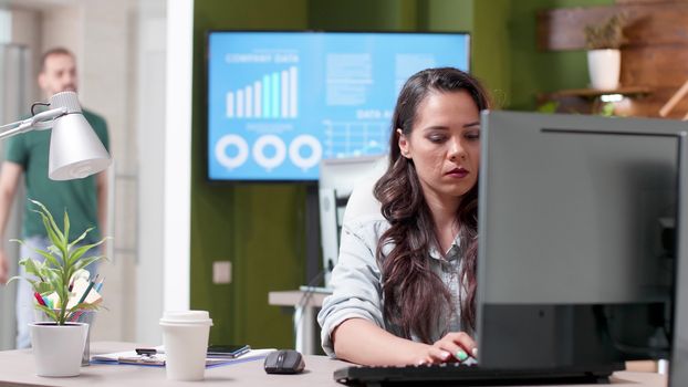 Revealing shot of a busy office with people working in it. A woman in casual clothes sips coffee and types on the computer