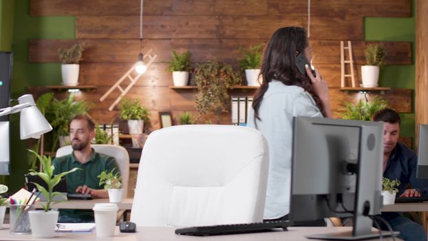 Female manager talks on the phone while walking in the office. Two colleagues are working on the computers in the background