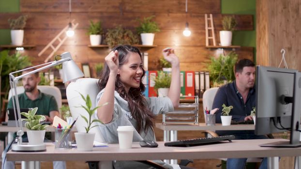 Female manager dances happily at her desk while her colleagues are working in the background. Slow motion medium shot