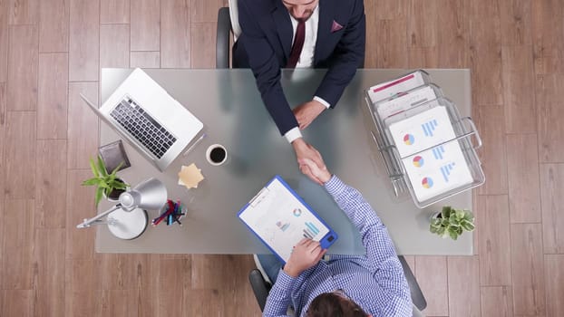 Two businessmen shaking hands sitting at the office with charts on clipboard, top view
