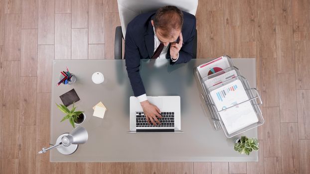 Top view of businessman in suit talking on the phone and typing on laptop sitting at his desk in the office.