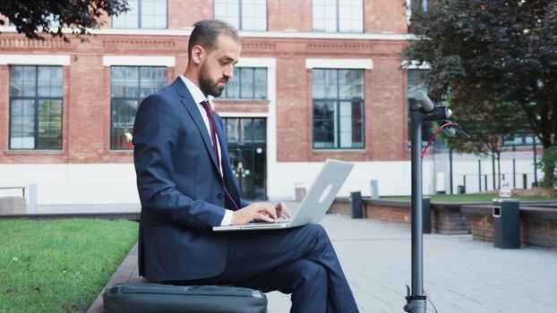 Man in suit sitting in business district working on the laptop. Next to his is an electric scooter