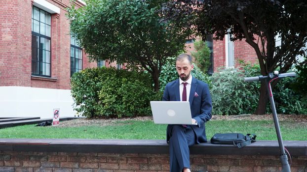 Manager in business suit working outside on the laptop. He has an electric scooter next to him. Slow motion footage
