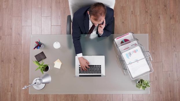 Top view of businessman in suit talking on the phone and typing on laptop sitting at his desk in the office.