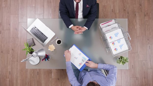 Top view of businessman in suit shaking hands with manager in his office. Clipboard with charts.