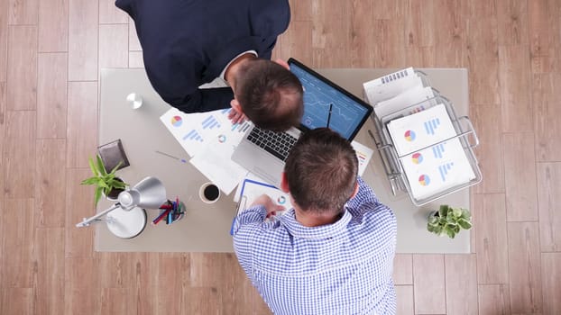Top view of businessmen working together and analysing charts from laptop in corporate office.