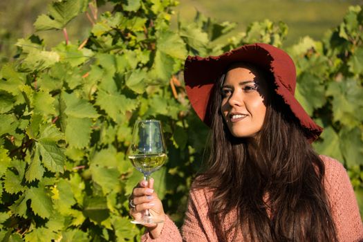 Outdoor portrait of gorgeous young female enjoying a glass of wine in a vineyard. Female with staylish hat in vineyard.