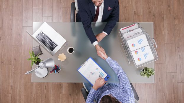 Two businessmen shaking hands sitting at the office with charts on clipboard, top view