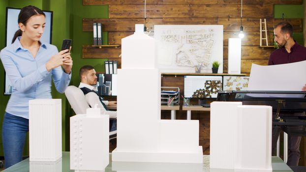 Young female architect taking photos of building models. Bearded architects working in the background.