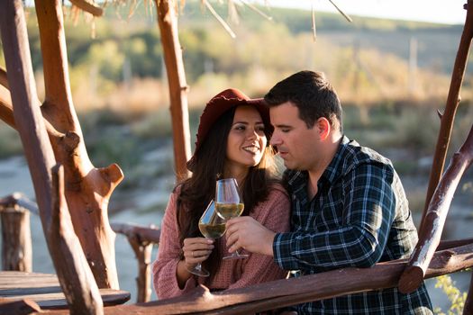 Happy young wife looking at her husband while enjoying together a glass of wine by the edge of a lake in the middle of a vineyard. Beautiful young couple.