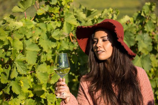 Beautiful young female with stylish hat in a vineyard holding a glass of white wine. Portrait of beautiful brunette in the vineyard.