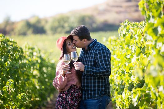 Attractive young boyfriend kissing his gorgeous wife on the rows of green vineyard. Adorable couple between rows of vine.