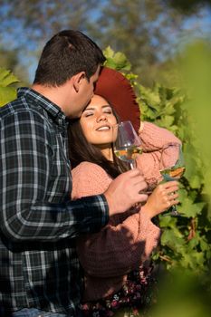 Husband kissing his wife forehead in a green vineyard and holding a glass of wine.