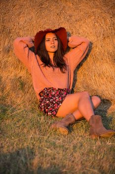 Happy young woman with stylish hat at sunset with big straw bale behind it.