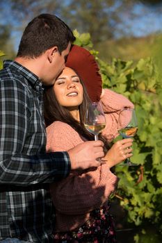 Happy young wife with a stylish hat a in vineyard on the country side. Husband kissing his wife in direct sunlight.