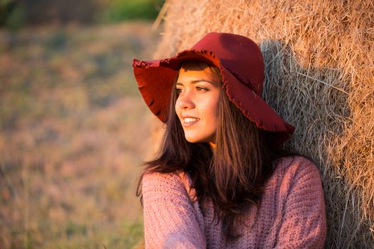 Side view portrait of beautiful young woman with stylish hat at sunset light with vineyard and straw bale behind her.