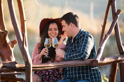 Couple sitting on a wooden pontont at the edge of a lake in the middle of a vineyard dinking a glass of wine.