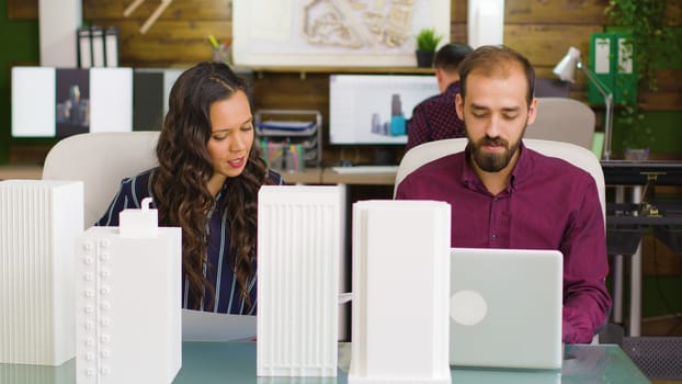 Designer working on a laptop and architec looking at blueprints in modern office interior