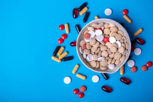 Top view with a bowl of multi-colored pills and next to other pills of different colors and shapes. Copy space and blue background