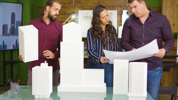 Team of architects working with blueprints and 3D printed models of buildings in modern cozy office
