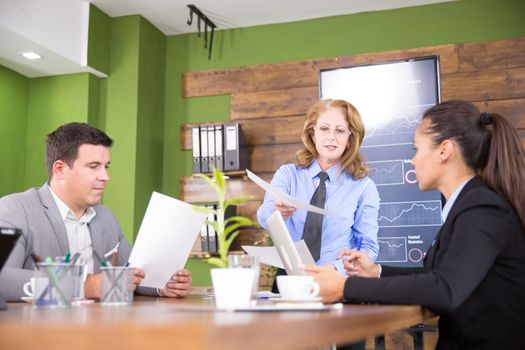 Executive female giving her assistant charts in a conference room. Tv screen with charts.