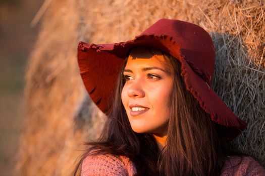 Smiling young brunette with straw bale behind her at sunset.