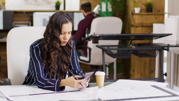 Blueprints on professional architect table looking at her phone.
