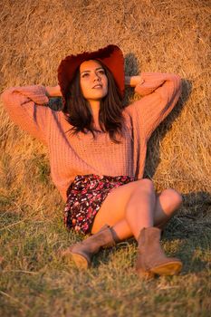 Attractive young woman with stylish hat looking up. Woman at sunset with straw bale behind her.