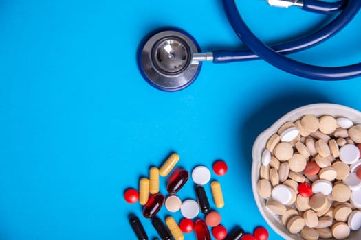 Top view with a stethoscope and pills of many colors and a bowl of pills. Blue background and copy space