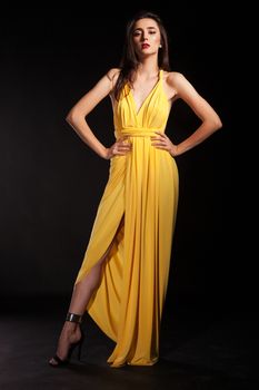 Stylish brunette female wearing yellow fashion dress on black background holding hands on her hips.