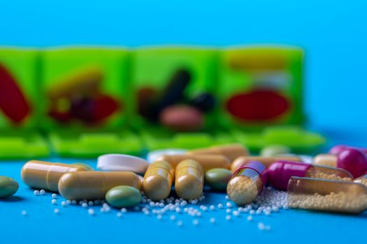 Top view with multicolored pills and capsules and in the back you see an organizer. Green organizer and blue background