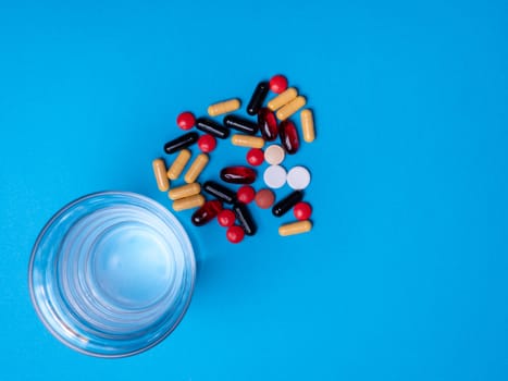 Top view with a glass of water and next to it multicolored pills and capsules. Copu space and blue background