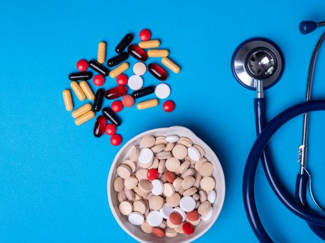 Top view with multicolored pills and capsules next to a stethoscope and a pill bowl. Blue background and copy space