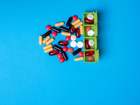 Top view with a green organizer with pills in it and next to multicolored pills and capsules . Blue background and copy space