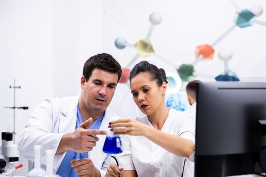 Scientist in white coat and his assitant looking at the bottle with blue liquid in modern research laboratory.