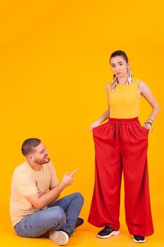 Boyfriend pointing at her beautiful girlfriend funny pants in studio over yellow background.