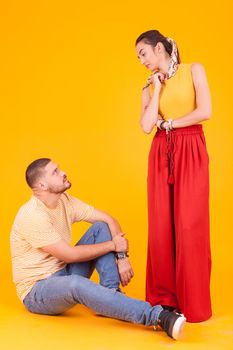 Confident boyfriend looking at his beautiful girlfriend sitting down over yellow background.