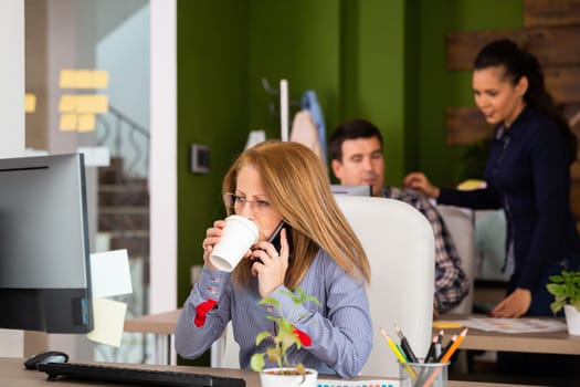 Mature business woman talking on the phone and taking a sip of coffee. Assistants working in the background.