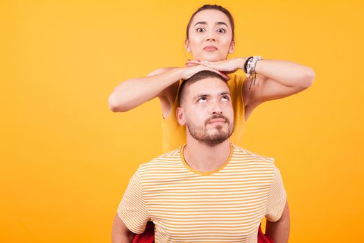 Beautiful couple having fun while man piggybacking his girlfriend in studio over yellow background.