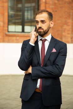 Young man dressed in a suit talking on the phone near a building. Success and communication