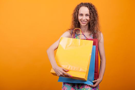 Cute woman in stylish dress holding colorful shopping bags over orange background in studio.