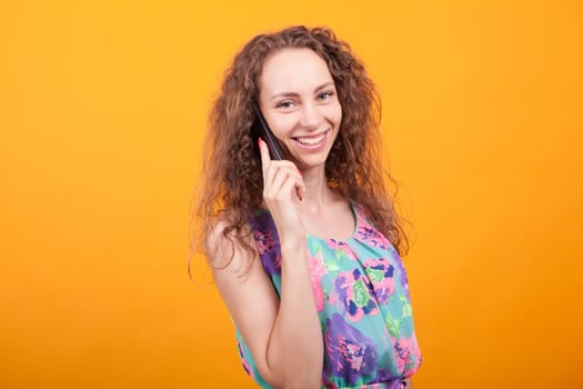 Beautiful young woman in summer dress talking on the phone smiling over orange background in studio.