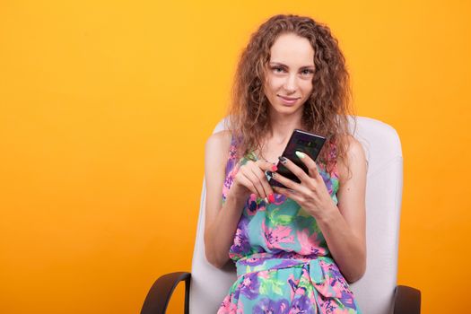 Woman in summer dress smiling to the camera and holding her mobile phone in studio over orange background.