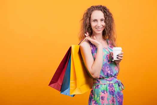 Smiling woman holding her shopping bags and her tasty coffee in studio over orange background.