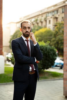Young businessman dressed in suit talks on the cellphone on the street. Sunset and street background