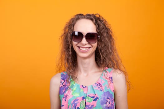 Portrait of young woman smiling and wearing sunglasses in studio over orange background. Woman in trendy dress.