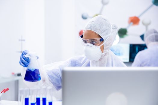 Researcher looks at a blue liquid container in front of a laptop. Glasses and protective suit