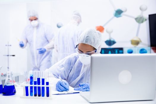 Researcher notes something on the sheet while she has several tubes and a laptop in front of her. Behind her two colleagues work