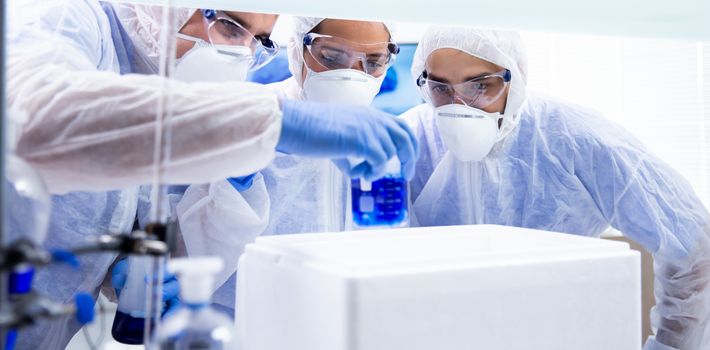 Three chemists look at a beaker with a blue substance in it. Laboratory and white box